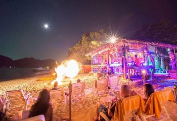 Phi Phi Hotel beachfront bar at night. Gay-friendly resort with private beach. People relaxing by firelight. Tropical travel.