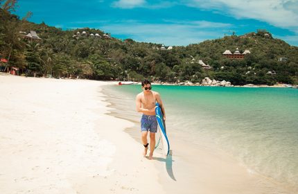 Gay traveler at Anantara Rasananda Koh Phangan Villas. Man walks on beach with board. Luxury beach resort for gay travelers. Thailand travel.