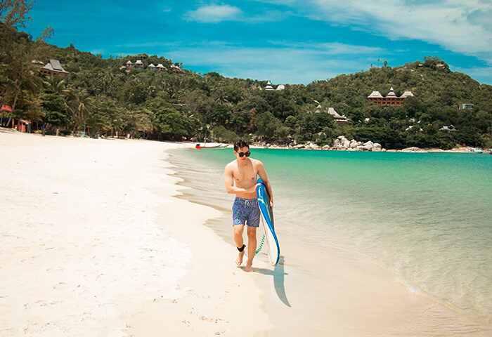Gay traveler at Anantara Rasananda Koh Phangan Villas. Man walks on beach with board. Luxury beach resort for gay travelers. Thailand travel.