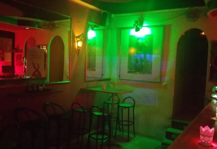 Interior of Chessa Bar, a gay bar in Torremolinos, Spain. Green and red lights illuminate the bar area with stools and artwork on the walls.