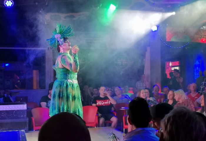 Chez Funny Boys gay bar in Gran Canaria: Drag queen in green gown performs on stage for a seated audience, stage lights, smoke.