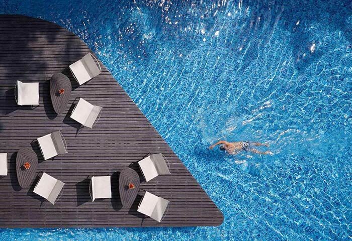 Baraquda Pattaya Mgallery: Chic design gay hotel in Pattaya with pool. Man swims in clear blue water near lounge chairs on a wooden deck.