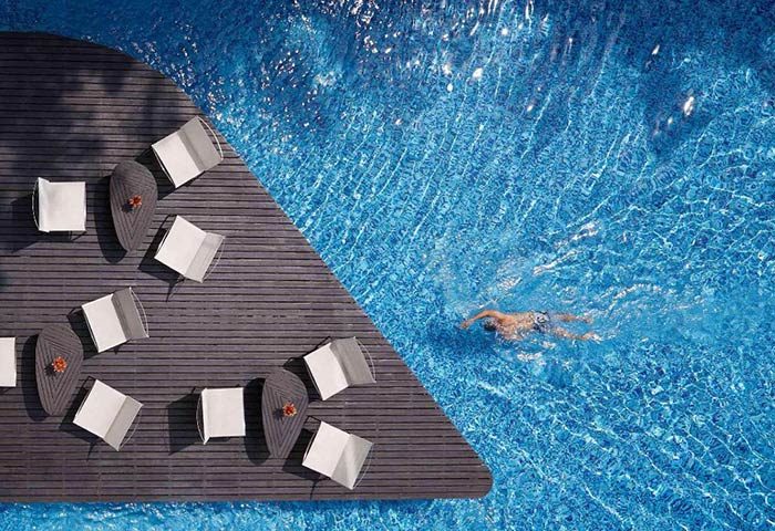 Baraquda Pattaya Mgallery: Chic design gay hotel in Pattaya with pool. Man swims in clear blue water near lounge chairs on a wooden deck.