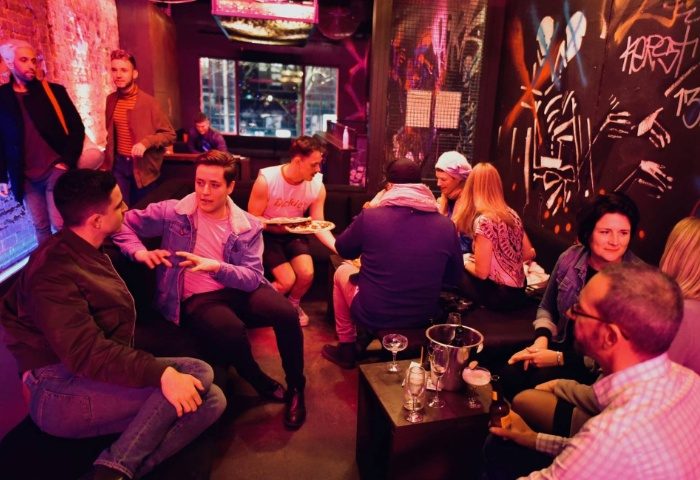 Ching-a-Lings Sydney: Gay men socializing at a bar with graffiti art. People are drinking and chatting in a dimly lit, trendy space.