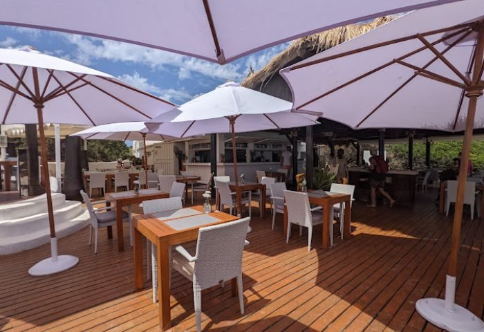 Outdoor seating at Chiringay gay bar and beach in Ibiza. Tables and chairs under umbrellas on a sunny day. Gay travel destination.