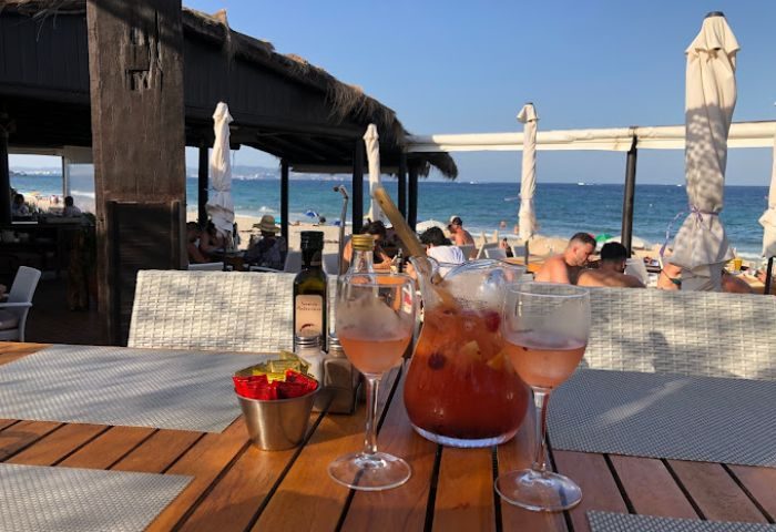 Chiringay Gya Bar, Ibiza beach. Refreshments on a table, with beachgoers and the sea in the background. Gay travel destination.