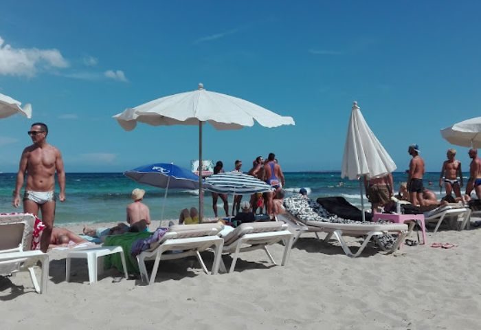 Ibiza's Chiringay Gya Bar beach scene: Gay men relax on lounge chairs under umbrellas, enjoying the sun and sea.