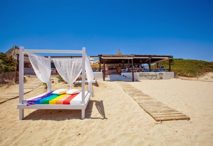 Chiringay gay bar and beach in Ibiza. Beach bed with rainbow flag and white curtains. Wooden path to the bar. Gay travel advice.