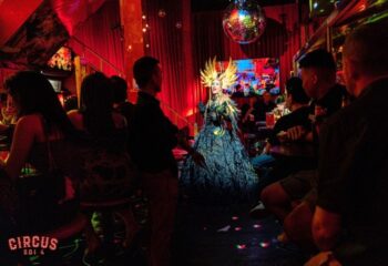 Circus Soi 4 Gay Bar in Bangkok: Drag queen performing in a dark, red-lit bar with a disco ball, entertaining a crowd of gay men.