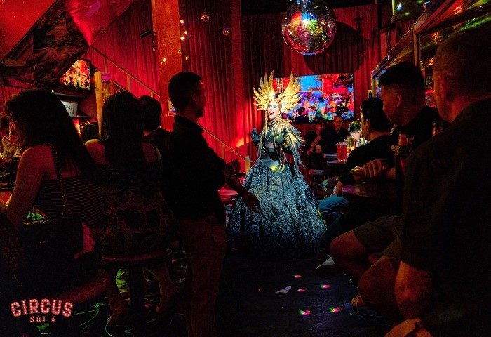 Circus Soi 4 Gay Bar in Bangkok: Drag queen performing in a dark, red-lit bar with a disco ball, entertaining a crowd of gay men.