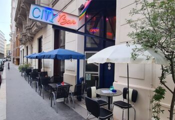 Vienna City Bar exterior. Gay bar with rainbow flag, tables, and umbrellas on the sidewalk. Gay travel in Vienna.