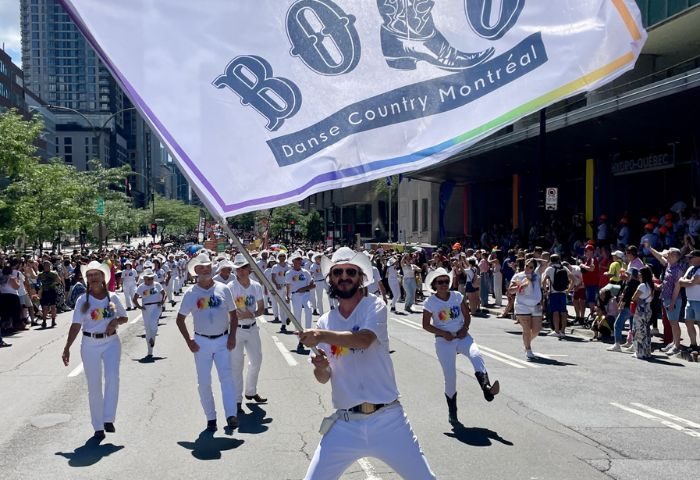 Montreal Gay Dance Party: Bolo Club dancers in white outfits and cowboy hats parade with a rainbow flag during Montreal Pride.