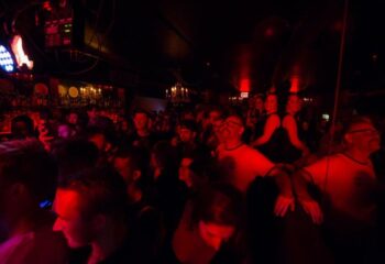 Inside Club Cumming in New York, a lively gay bar. A crowd of gay men and others gather under dim, red lighting and bar fixtures.