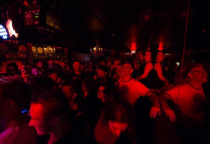 Inside Club Cumming in New York, a lively gay bar. A crowd of gay men and others gather under dim, red lighting and bar fixtures.