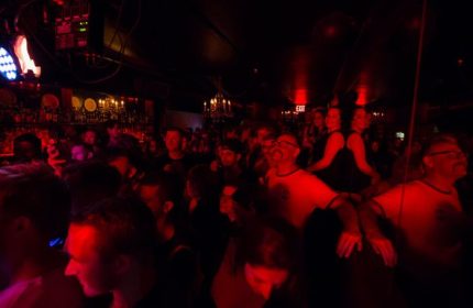 Inside Club Cumming in New York, a lively gay bar. A crowd of gay men and others gather under dim, red lighting and bar fixtures.