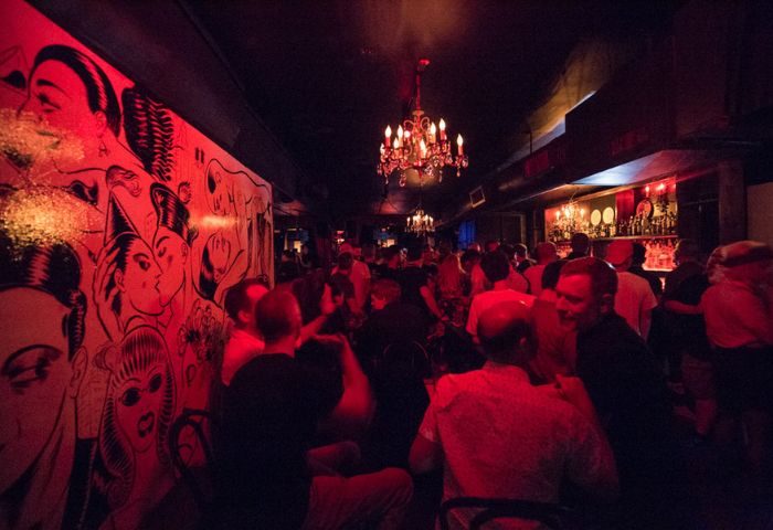 Inside Club Cumming in New York, a gay bar. Red lighting illuminates a crowd, a mural, and chandeliers. Gay travel advice.