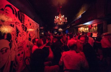 Inside Club Cumming in New York, a gay bar. Red lighting illuminates a crowd, a mural, and chandeliers. Gay travel advice.
