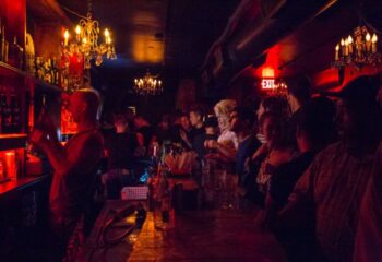 Inside Club Cumming in New York, a popular gay bar. Patrons stand at the bar, illuminated by chandeliers and red lighting. Bartender visible.