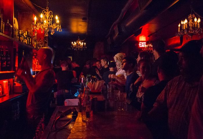 Inside Club Cumming in New York, a popular gay bar. Patrons stand at the bar, illuminated by chandeliers and red lighting. Bartender visible.