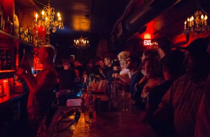 Inside Club Cumming in New York, a popular gay bar. Patrons stand at the bar, illuminated by chandeliers and red lighting. Bartender visible.