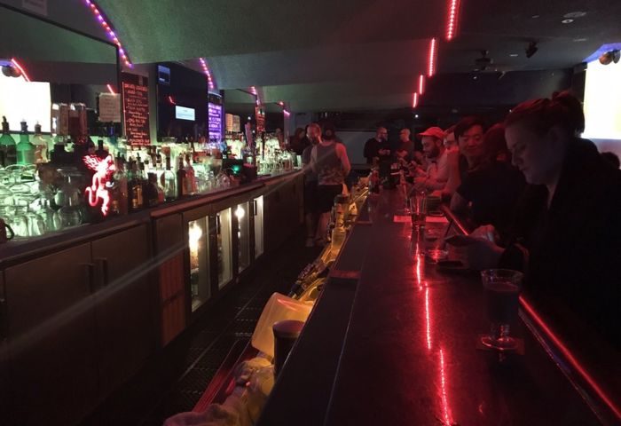 Interior of Club OMG, a gay bar in San Francisco. Patrons sit at the bar under red neon lights. Shelves of liquor bottles behind.