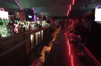 Interior of Club OMG, a gay bar in San Francisco. Patrons sit at the bar under red neon lights. Shelves of liquor bottles behind.