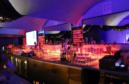 OMG Gay Bar in San Francisco: A long bar with bottles, glasses, and drink menus illuminated with red and blue lights. Nightlife.