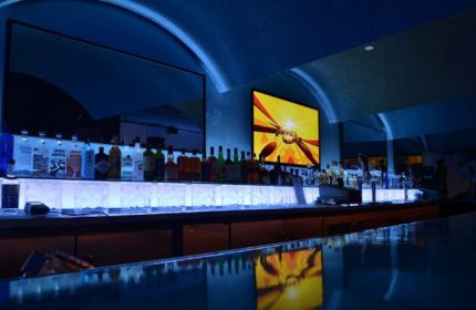 Interior of Club OMG, a gay bar in San Francisco. Bottles line the bar with blue lighting and a screen showing abstract art.