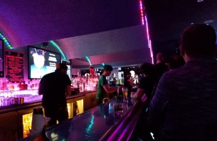 Inside Club OMG, a gay bar in San Francisco. Bartender serving drinks, patrons at the bar, purple lighting. Gay travel advice.
