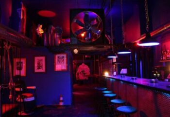 Interior of Open Mind gay cruise club in Barcelona. Bar with stools, industrial decor, red and blue lighting, and adult-themed art.