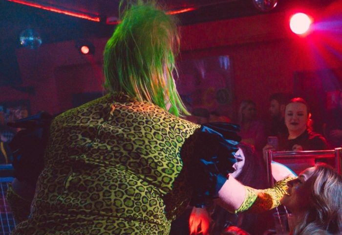 Club Tropicana Manchester scene. Drag queen with green hair in leopard print dress interacts with a smiling woman in a gay club.