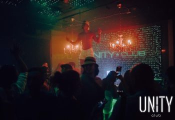 Gay party at Unity Club, Montreal. A shirtless dancer performs above the crowd. Chandeliers and club logo in the background. Nightlife.