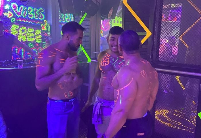 Three shirtless men with neon body paint at Club XXL, a gay club in Sitges. Nightlife scene, vibrant and fun atmosphere. Gay travel.