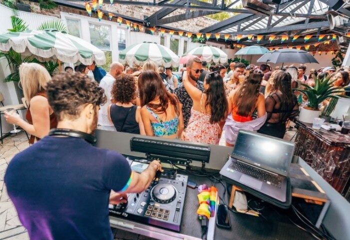 DJ at Colombian Hotel Sydney LGBTQ+ event. Crowd dancing under rainbow flags & umbrellas. Gay travel party scene.