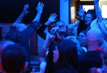 Crowd at Coming Out Bar, Rome, raises hands in excitement. Drag queen in sequined top visible. Gay travel nightlife scene.