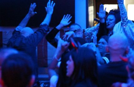 Crowd at Coming Out Bar, Rome, raises hands in excitement. Drag queen in sequined top visible. Gay travel nightlife scene.