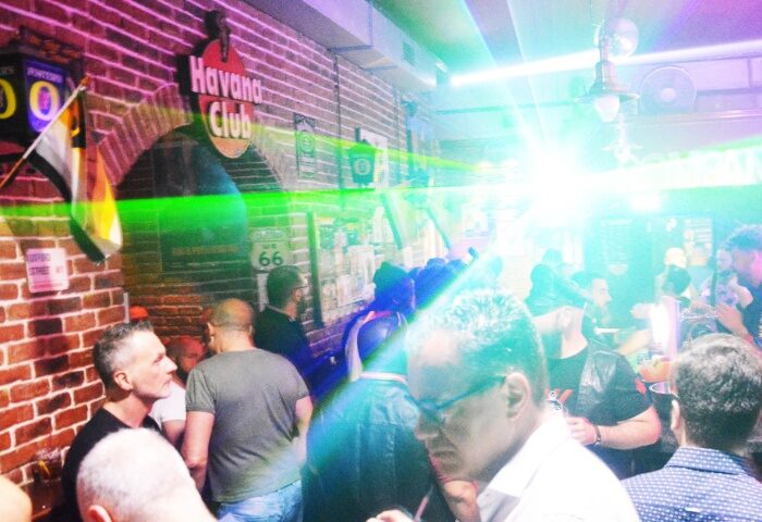 Company Club Milano: Gay men inside a club with a Havana Club sign on a brick wall, colorful lights, and a crowded dance floor.