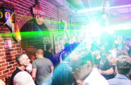 Company Club Milano: Gay men inside a club with a Havana Club sign on a brick wall, colorful lights, and a crowded dance floor.