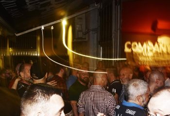 Company Club Milano crowd scene. Gay bar nightlife in Italy. Men socializing, blurred lights, and a lively atmosphere. Gay travel advice.