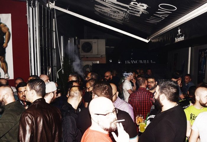 Crowded Company Club Milano gay bar scene. Men socializing, smoking, and drinking. Gay nightlife travel advice.
