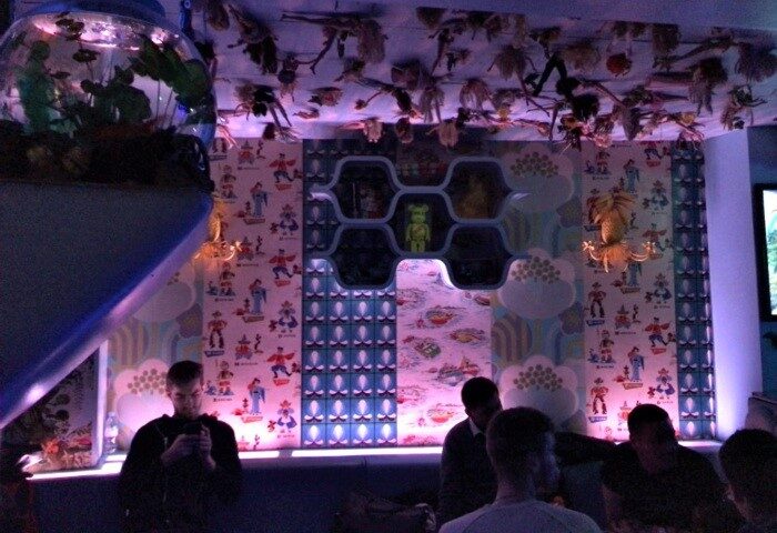 Cool Gay Bar London, The Friendly Society: Quirky interior with unique decor, including wallpaper, ceiling art, and a fish tank. Gay street scene.