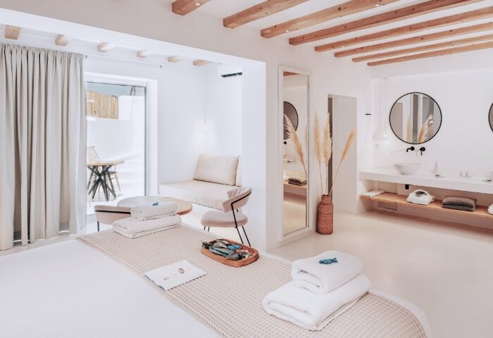 Akkadian Gay Hotel Mykonos design pool villa. Stylish room with white decor, wooden beams, towels, and bathroom. Gay travel luxury.