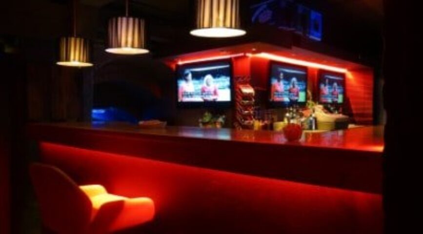 Covent Garden Health Spa's gay sauna bar area. Red lighting, TVs showing sports, and modern design. A popular spot for gay men's travel in London.