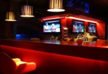 Covent Garden Health Spa's gay sauna bar area. Red lighting, TVs showing sports, and modern design. A popular spot for gay men's travel in London.