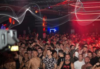 A packed gay party in Milan. Men crowd the dance floor under red lights and decorative white streamers. Nightlife scene for gay travel.