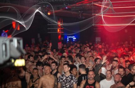 A packed gay party in Milan. Men crowd the dance floor under red lights and decorative white streamers. Nightlife scene for gay travel.