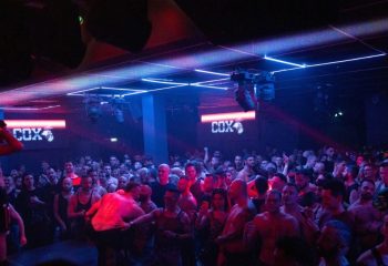Cox Party Milan: A crowded gay dance floor at a club, illuminated by blue and red lights. Men are dancing and enjoying the party.