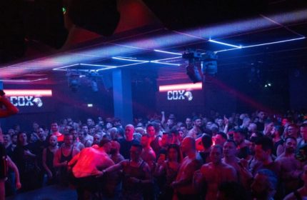Cox Party Milan: A crowded gay dance floor at a club, illuminated by blue and red lights. Men are dancing and enjoying the party.
