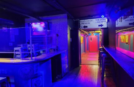 Interior of Crews & Tangos gay bar, Toronto. Hallway with bar, coat check sign, and colorful lighting. Gay travel advice.
