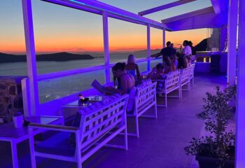 Crystal Cocktail Bar: Sunset view from a stylish bar. People enjoy drinks on white benches with blue cushions. Gay travel advice.